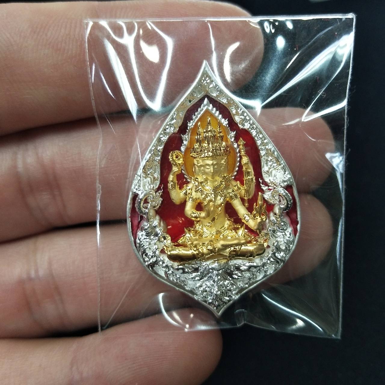 Thai wealth protection amulet gold deity in silver teardrop frame with red enamel held between fingers in plastic pouch
