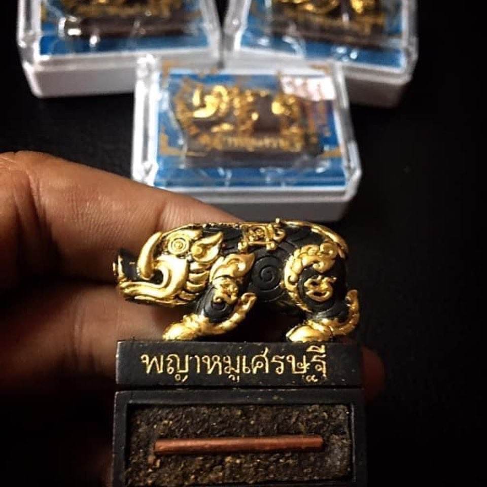 Hand holding Phaya Moo Setthi Amulet - Luang Por Whan’s gold and black pig talisman on pedestal with Thai script