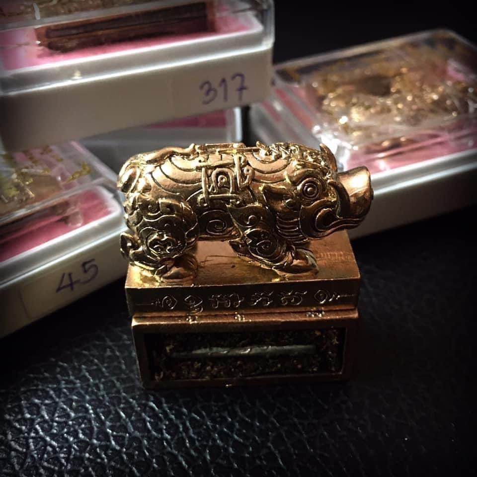 Gold pig Phaya Moo amulet on engraved pedestal with plastic case boxes in background Phaya Moo Setthi Amulet - Luang Por Whan’s.