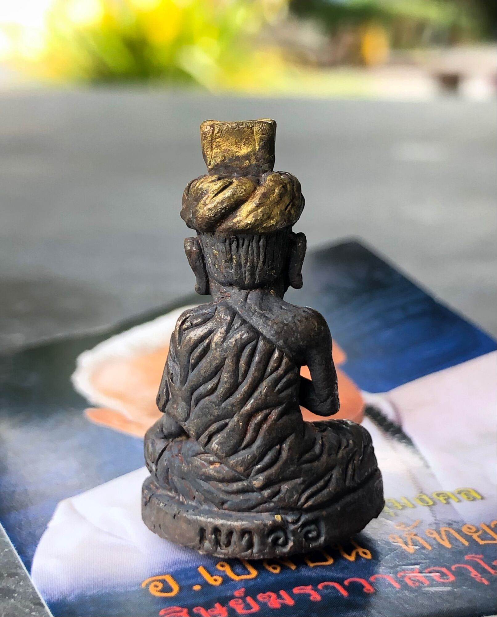 Bronze seated amulet showing detailed carved robe and base engraving, Borom Kru Phra Ruesi Ta Fai Amulet . placed on a brochure