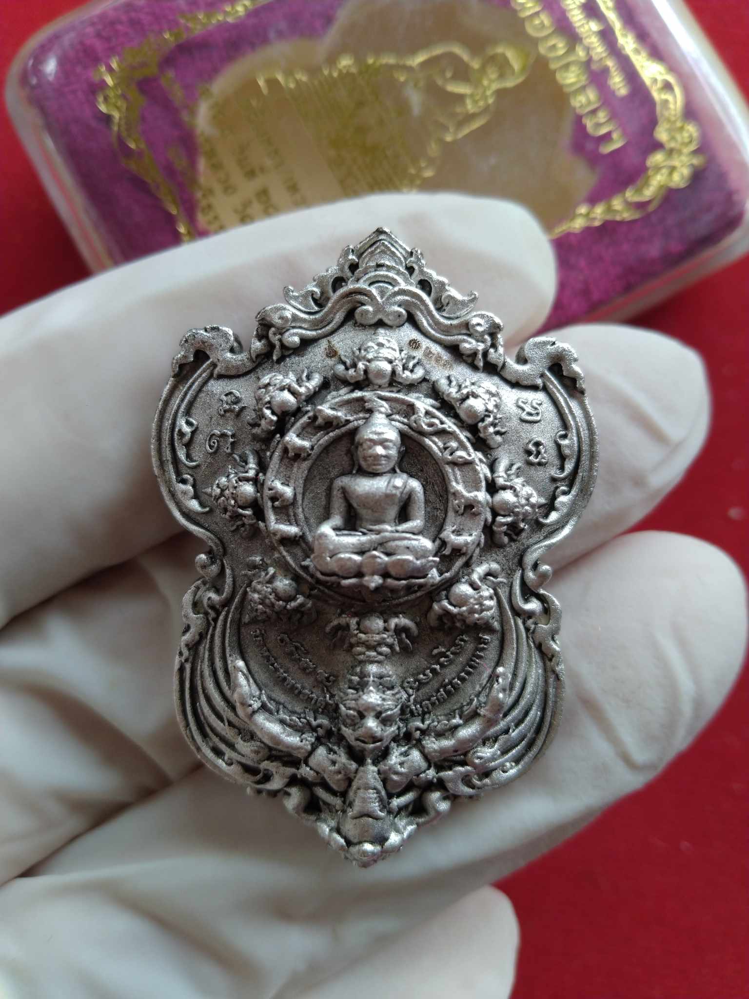 Gloved hand holding silver Sacred Phra Pidta Sri Maharaj Phangpakan. amulet pendant with seated Buddha and ornate mythical frame