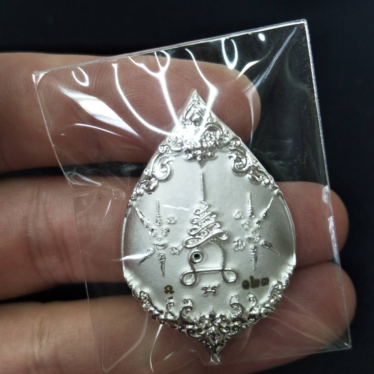 Thai wealth protection amulet silver teardrop pendant with ornate engraved designs in plastic pouch held by hand