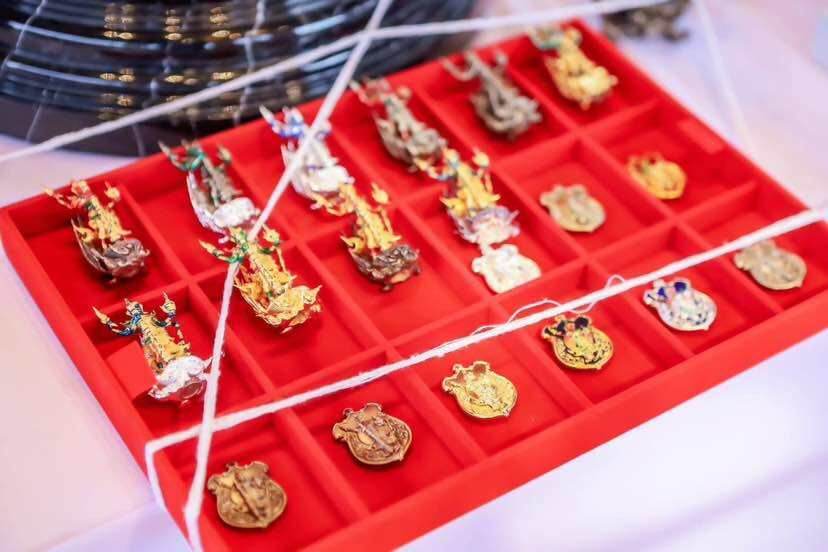 Red display tray of gold and enamel Thai amulet pendants in compartments secured by white string Thao Wessuwanno Sodaprom