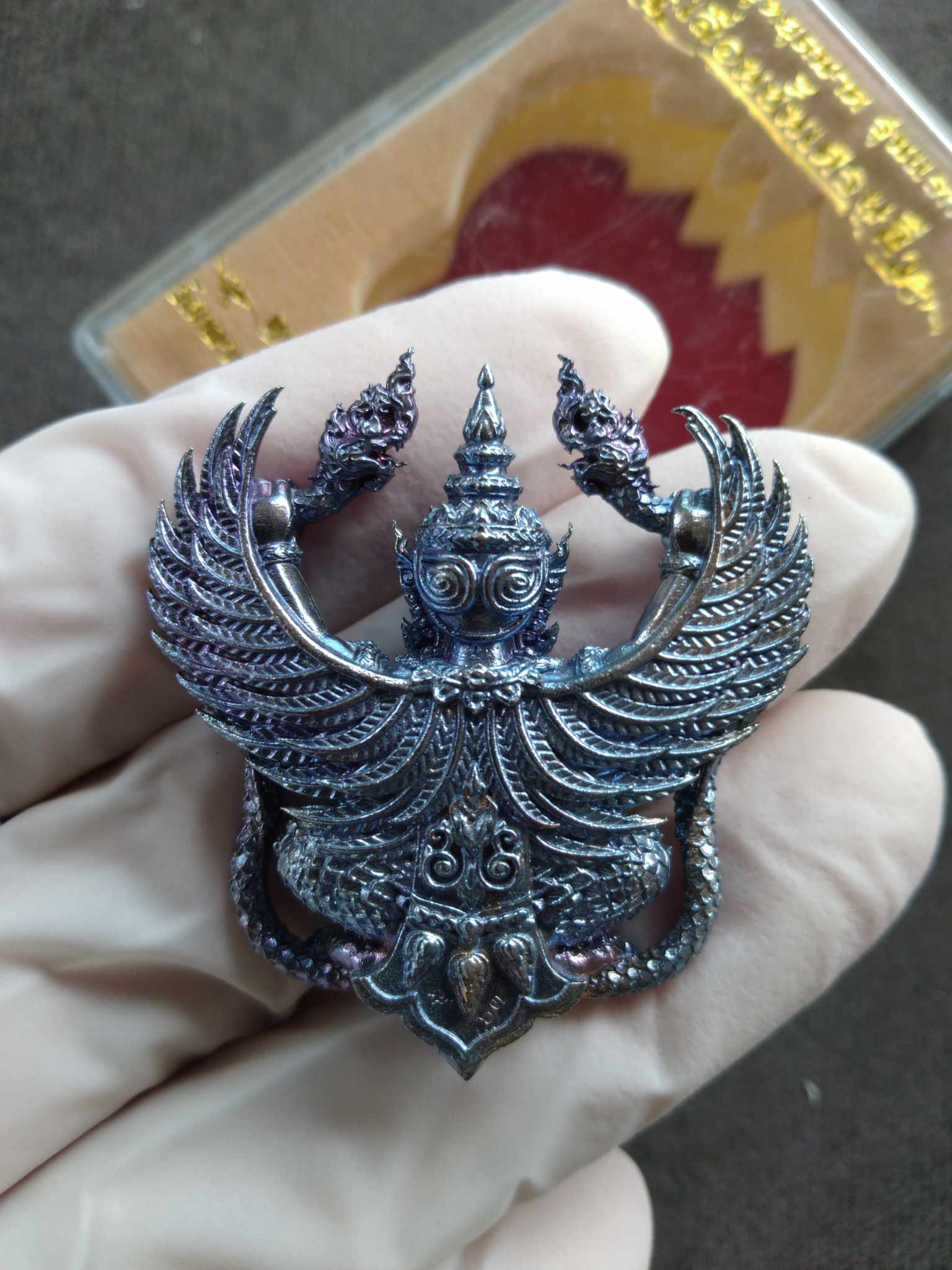 Hand holding detailed metal Garuda amulet blessed by Luang Pho Sanan. Intricate wings and ornate silver finish with display case