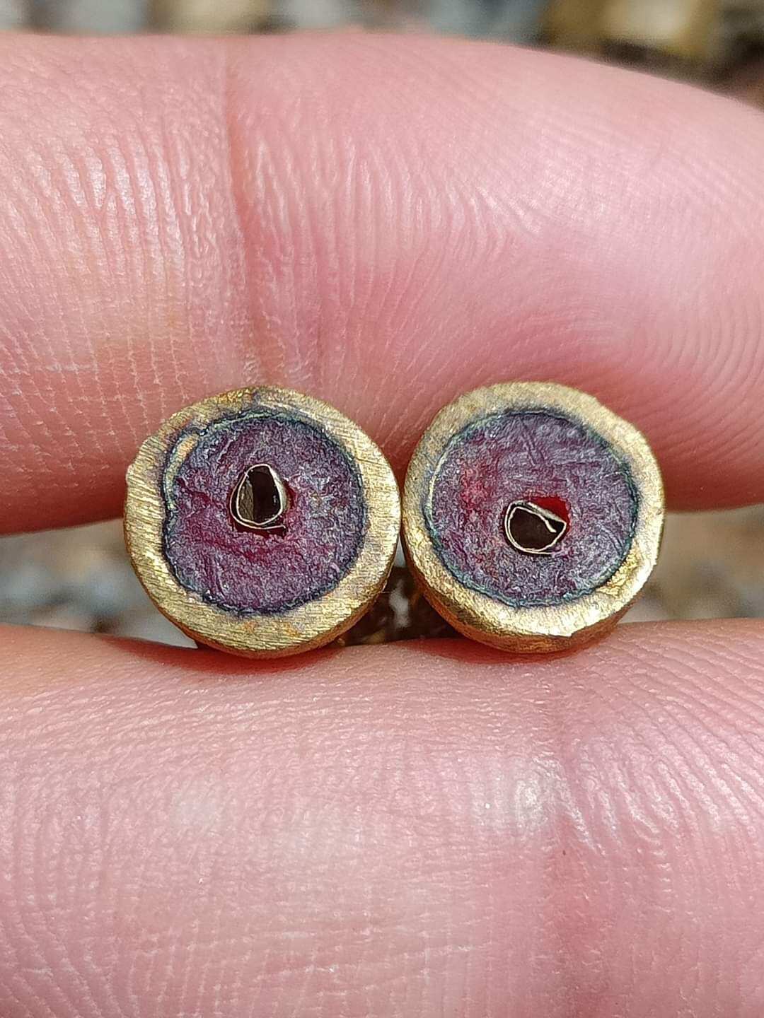 Close up of two small round Jeed Naree Phan Lak amulet pendants with gold rims and red centers held between fingers