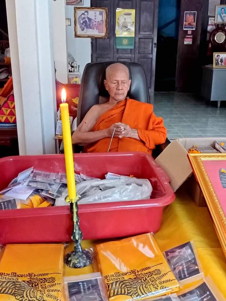 Luang Pu Thuad commemorative amulet 2023 display with monk seated behind table of packaged amulets and lit candle