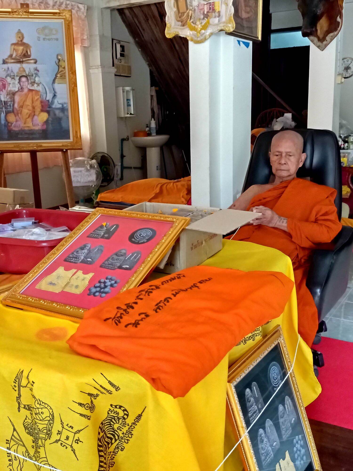 Thai Blessing Co display with monk and framed amulets featuring Luang Pu Thuad commemorative amulet 2023