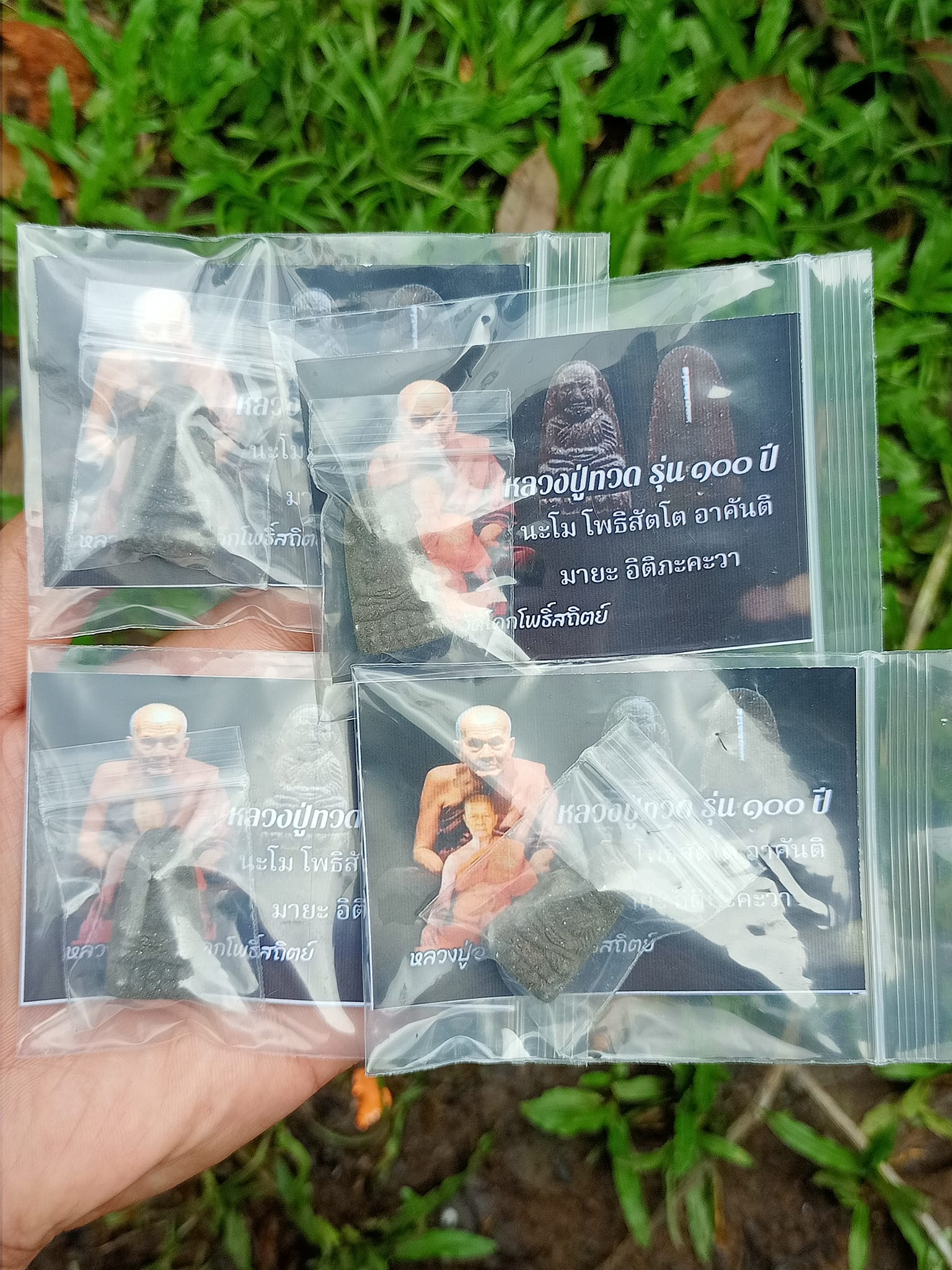 Hand holding four individually sealed Luang Pu Thuad commemorative amulet 2023 packages with display cards
