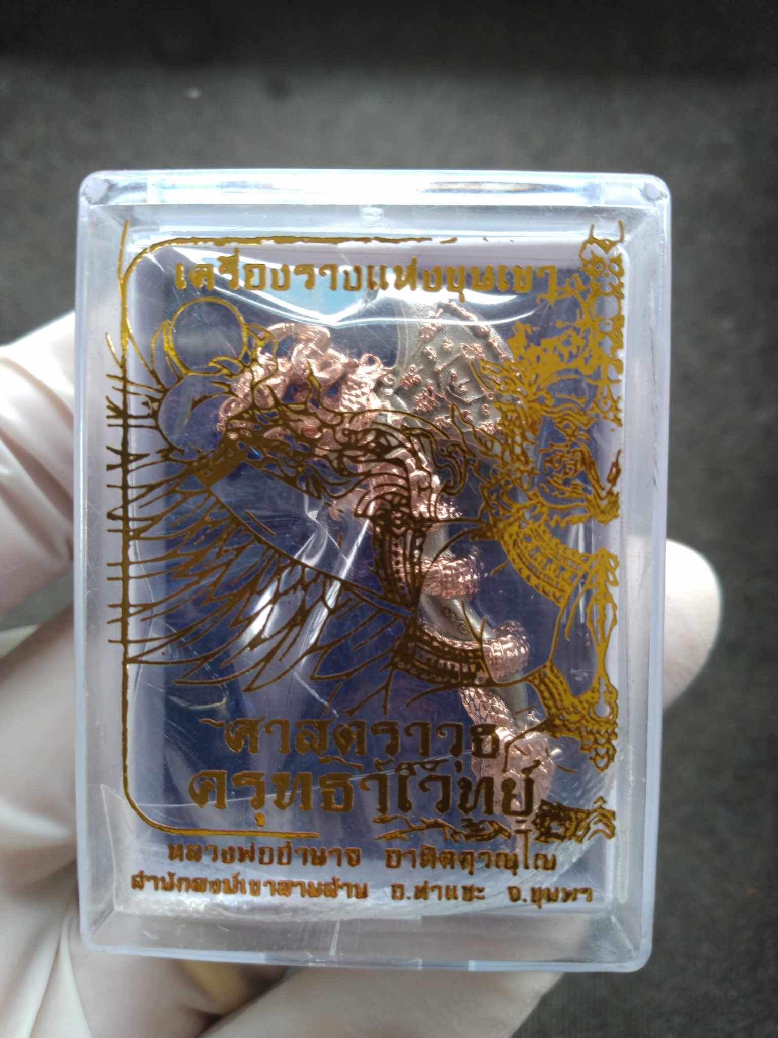 Sky Axe Krut Thawet amulet copper pendant in clear plastic case with gold Thai script held in gloved hand