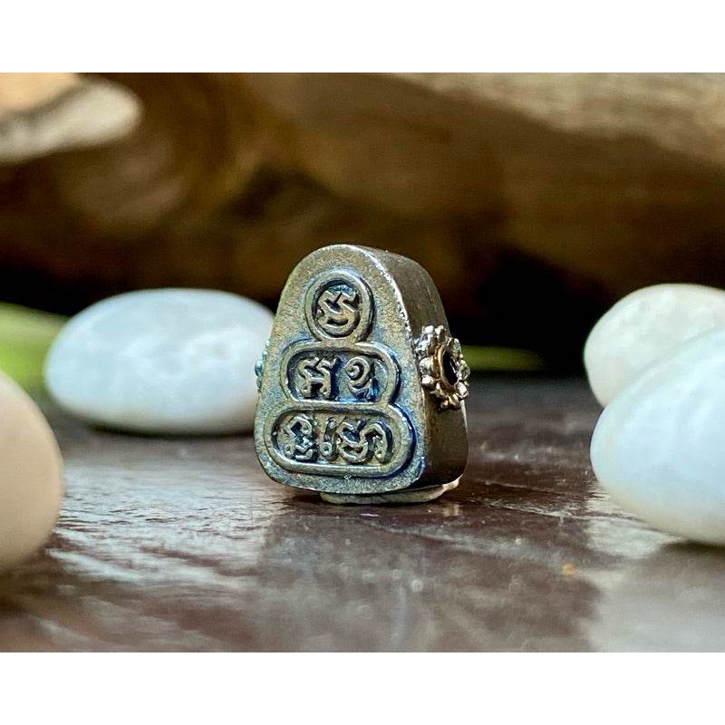 Thai billionaire wealth amulet small metal talisman with engraved sacred script and floral side detail on wood