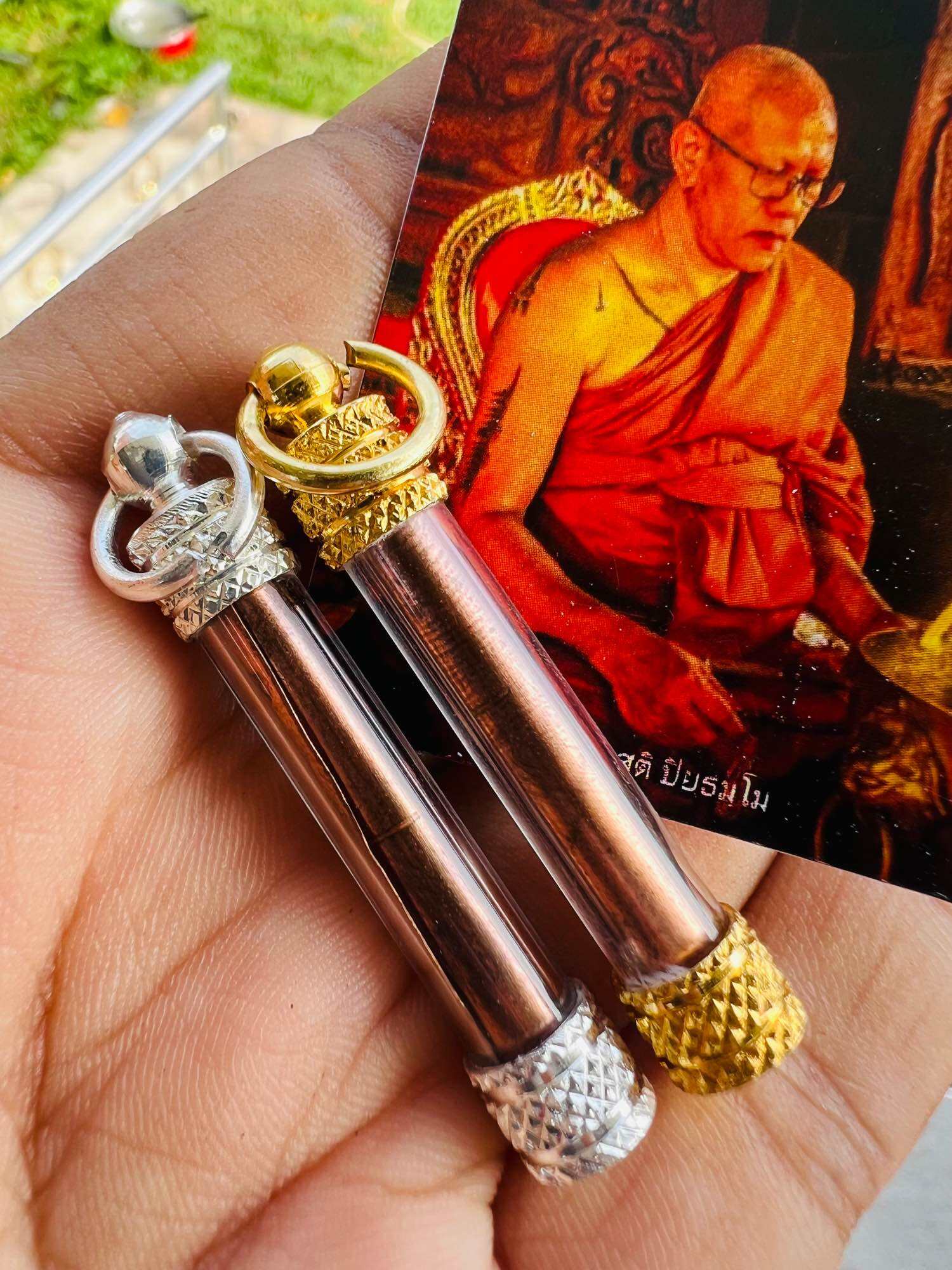 Hand holding two copper takrud tubes with gold and silver caps and rings and monk card Thai elephant takrud amulet