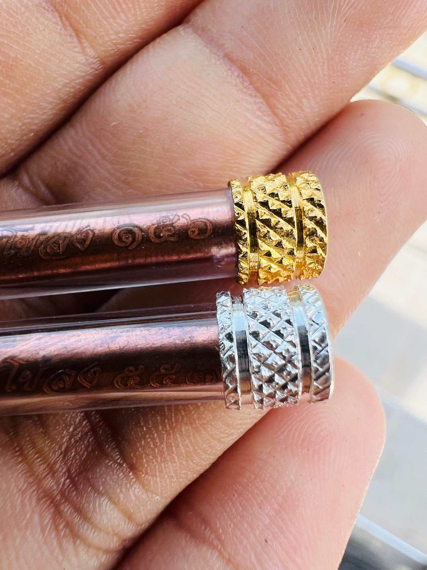 Hand holding two glass tube pendants with gold and silver caps revealing Thai elephant takrud amulet scrolls