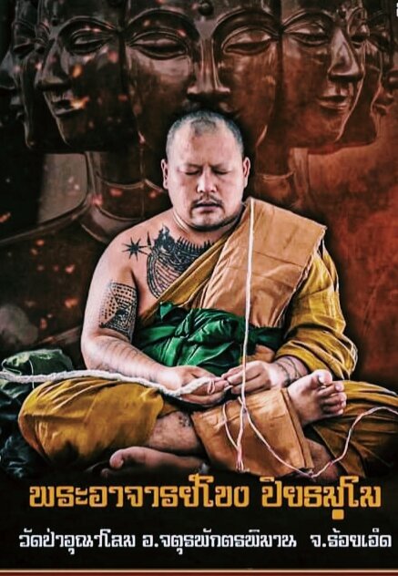 Tattooed meditating monk in saffron robes with carved deity backdrop for Thai guardian spirit protection charm