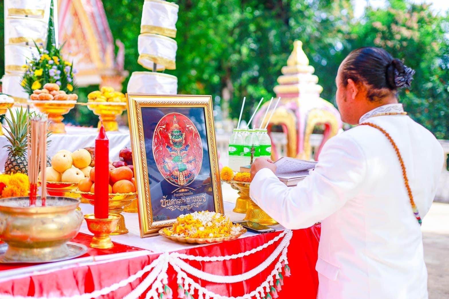 Thai ritual altar with framed deity image fruits candles and devotee in white making offering Thao Wessuwanno Bandarn Patthanapon