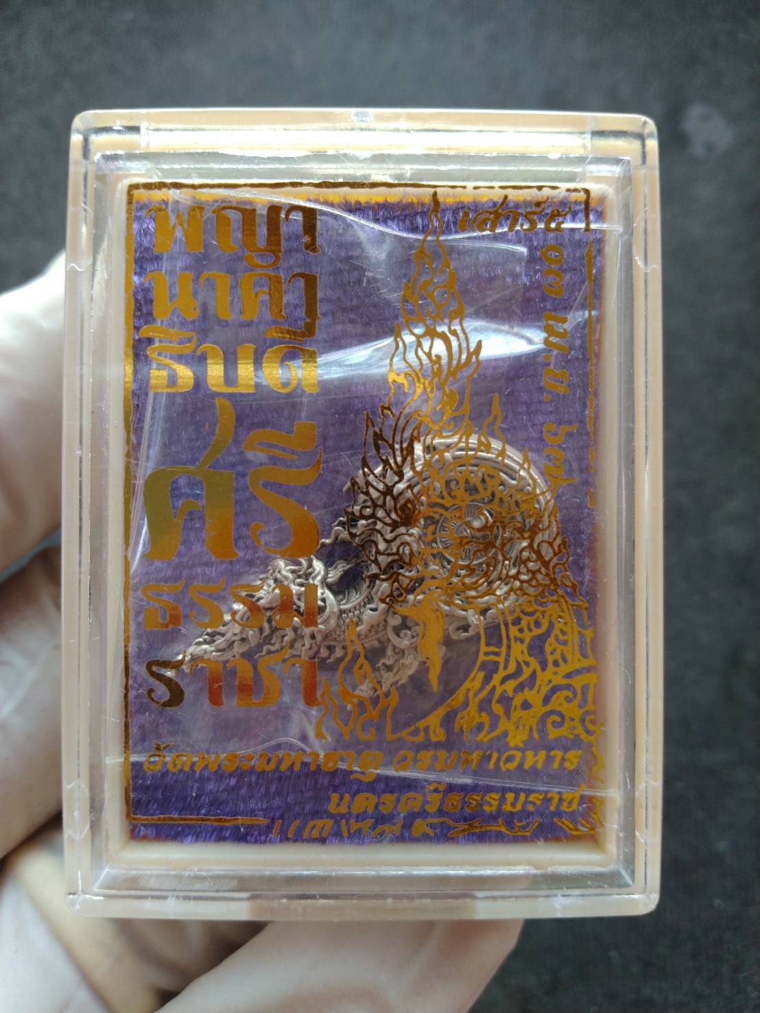 Phaya Naga amulet Thailand in clear plastic display case with gold foil design and purple background held by hand