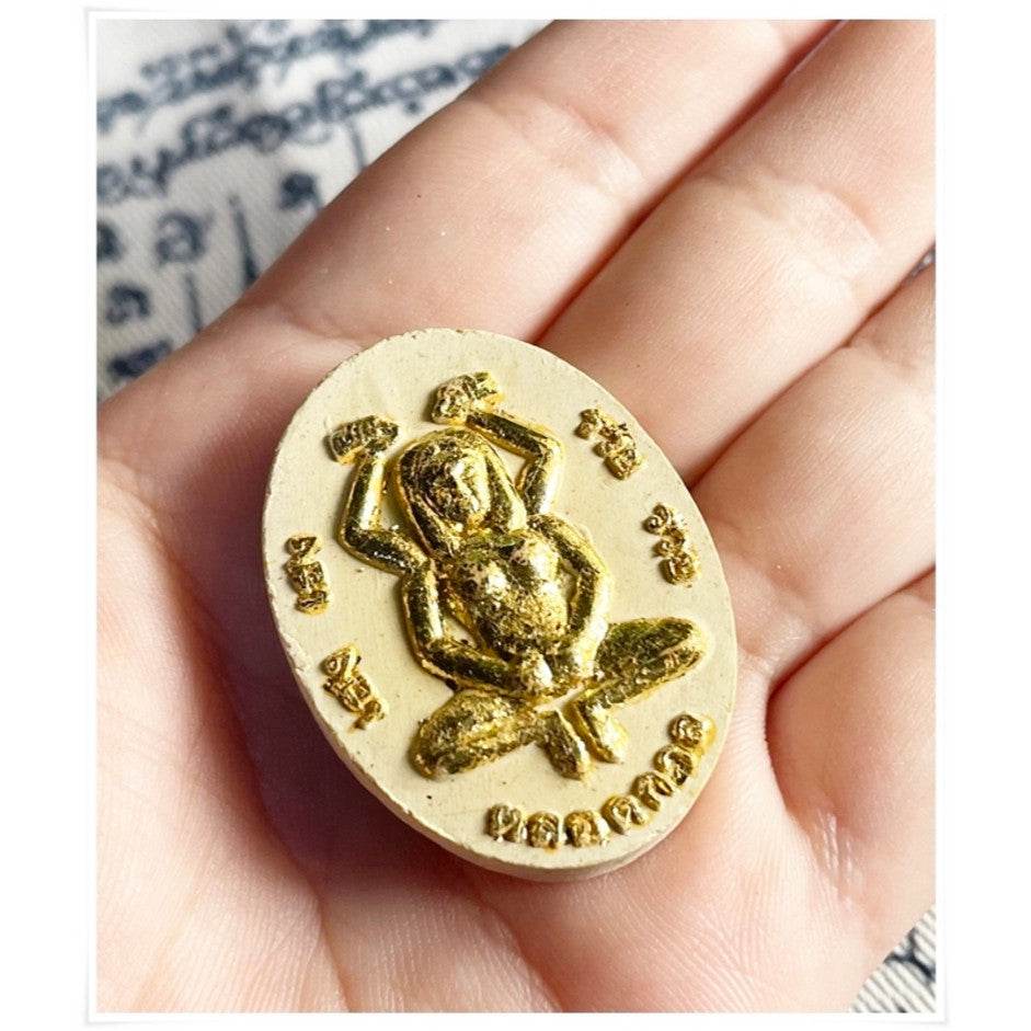 Hand holding a Divine attraction Thai charm with a golden frog design, symbolizing good luck and prosperity.