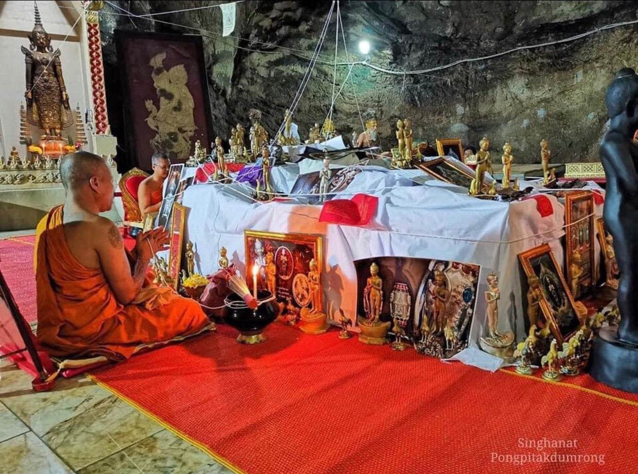 Monk praying near a display of Ei Khai Phaya Kai bronze amulet statues and framed images on a red carpet