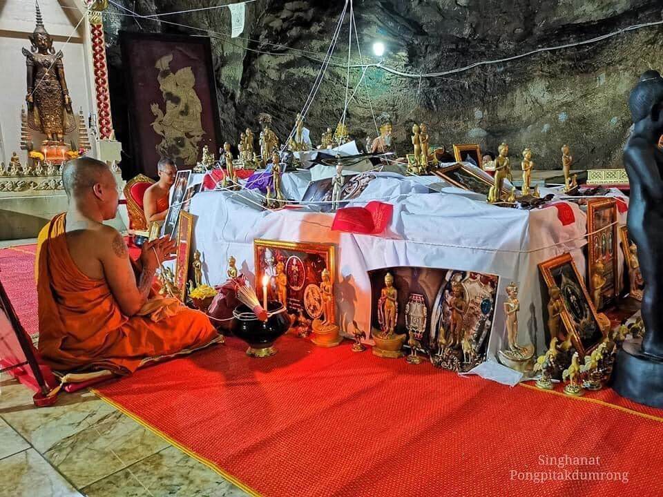 Monk praying beside an altar filled with Ei Khai Phaya Kai bronze amulet statues and framed images
