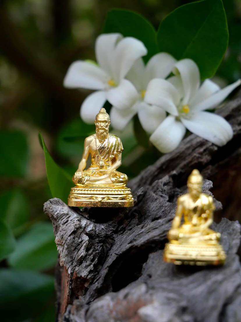 Gold Jivaka Komarabhacca amulet for health displayed on natural wood with white flowers