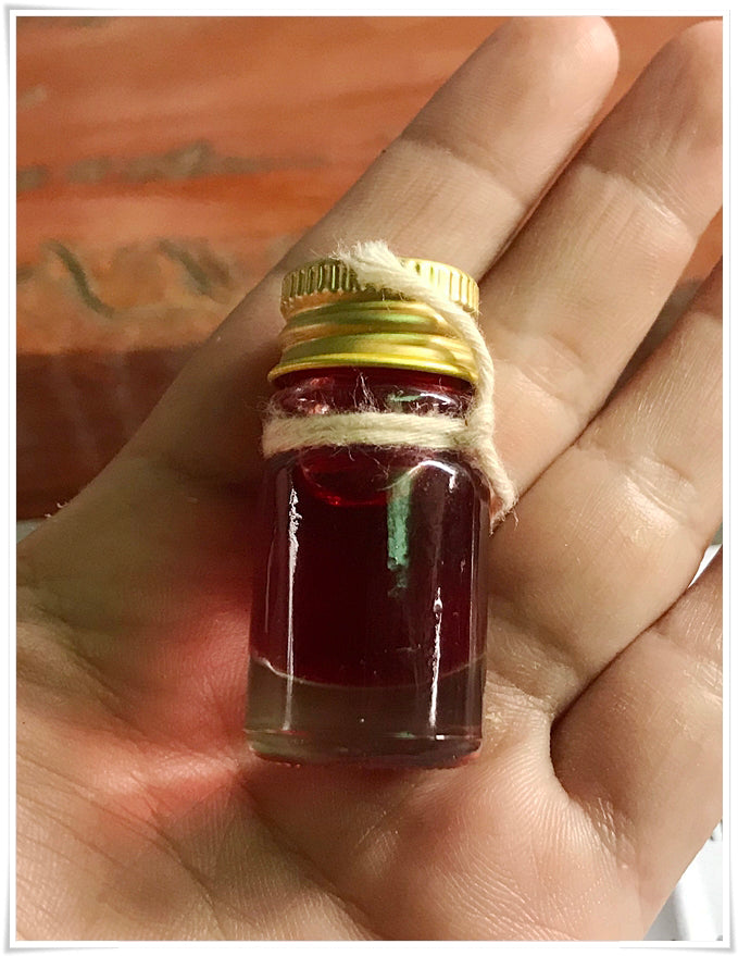 Small glass bottle of red oil in hand with gold cap and twine Lohit Sawat oil for attraction by Thai Blessing Co