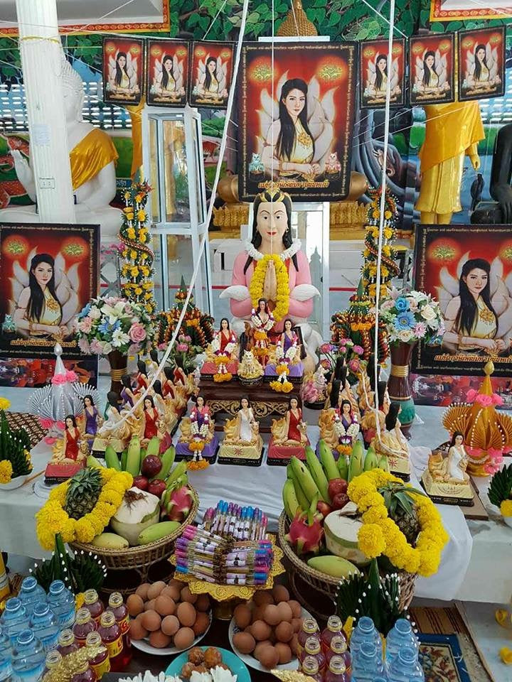 Altar display featuring Thai amulet for love and fortune with goddess statue garlands fruit eggs and bottled water