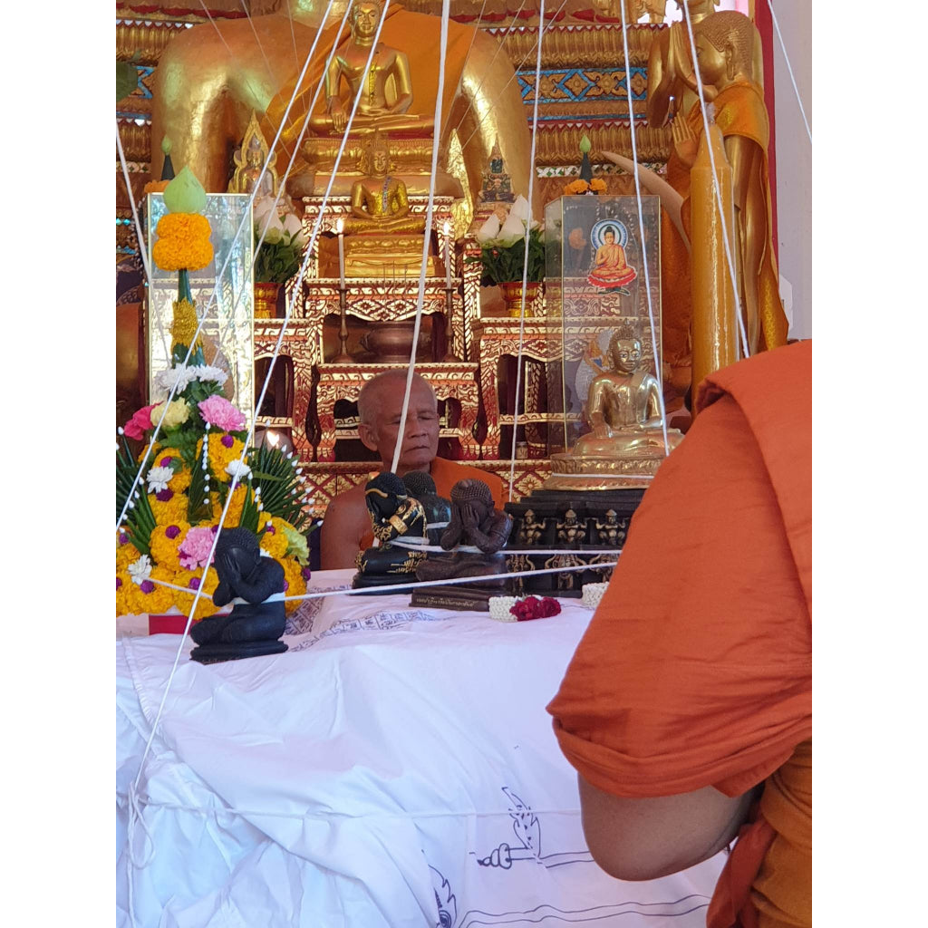 Monk blessing Phra Pidta amulet for wealth with traditional Thai Buddhist altar and floral arrangements