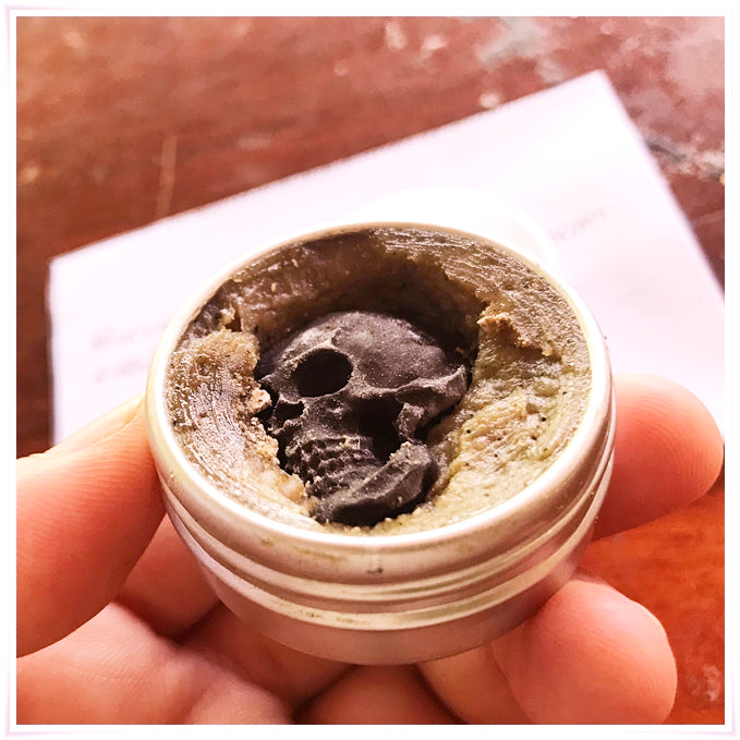 Hand holding small metal tin of herbal balm with carved black skull center Namman Prai balm for spiritual power by Thai Blessing Co