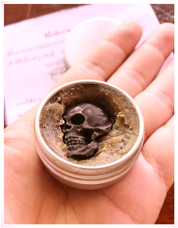 Hand holding Thai Blessing Co tin of Namman Prai balm for spiritual power showing green balm with skull amulet