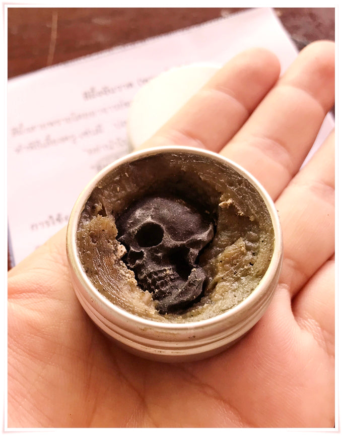 Hand holding open tin of Namman Prai balm for spiritual power showing skull shaped resin in green balm