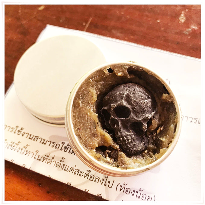 Tin of Namman Prai balm for spiritual power showing dark skull insert in herbal balm on paper