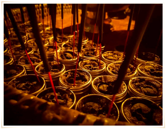 Rows of small tins of Namman Prai balm for spiritual power with incense sticks burning in warm temple lighting