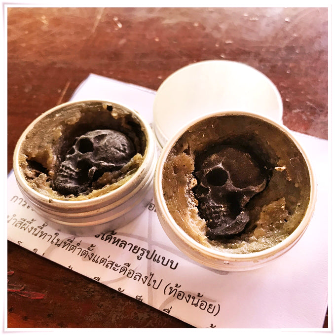 Two open tins of Namman Prai balm for spiritual power showing brown herbal paste with carved skulls