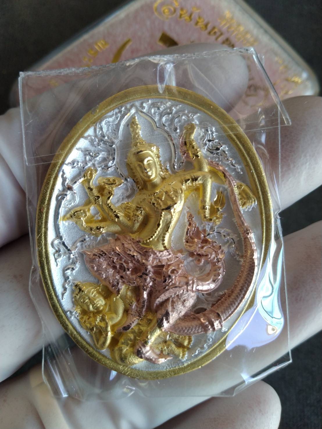 Hand holding authentic Thai Narai lucky charm gold silver copper oval deity pendant in plastic case