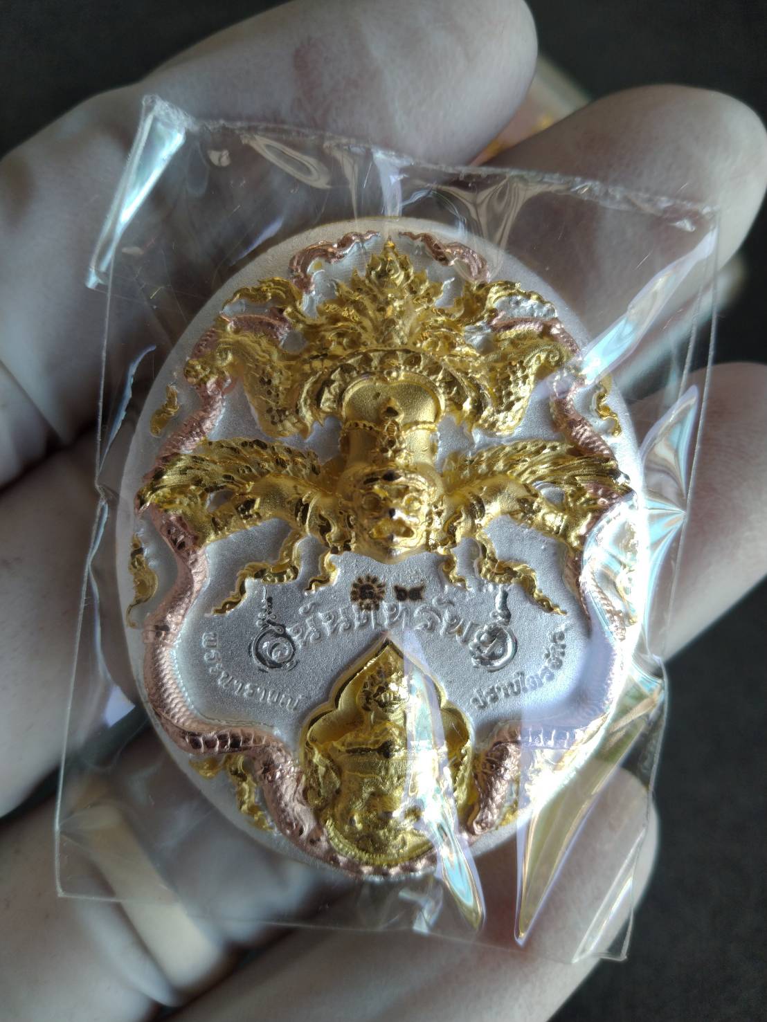 Hand holding authentic Thai Narai lucky charm gold and silver embossed amulet pendant sealed in plastic
