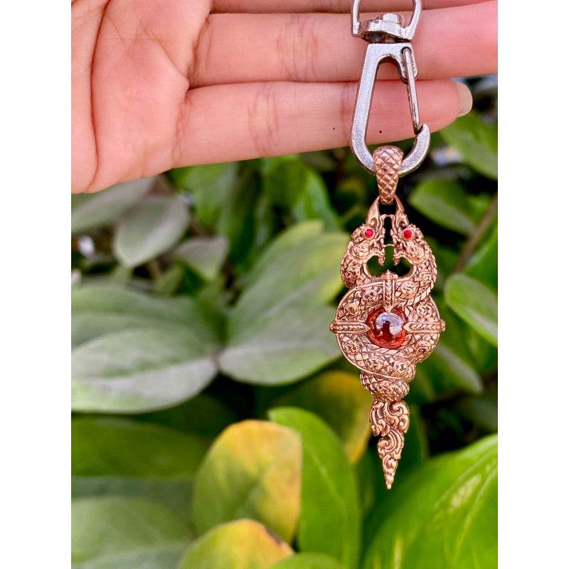 Thai Blessing Co Naga wealth elevation amulet keychain in gold tone with red gem held by hand over green leaves