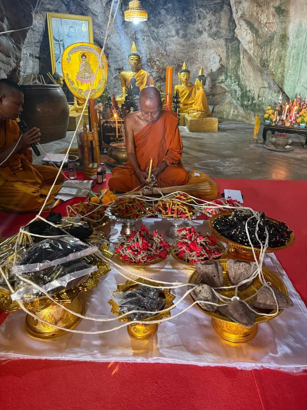 Monk performing ritual in cave temple blessing multiple amulet pendants on trays with cords blessed Thai Phan-Thai amulet for luck