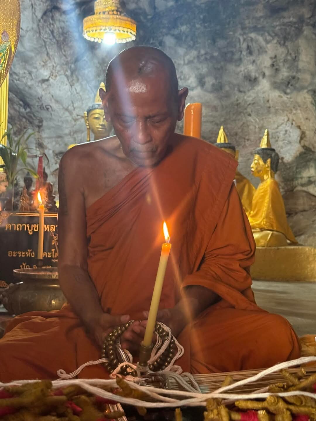 Monk blessing a pendant with candle and beads in temple cave holding blessed Thai Phan-Thai amulet for luck
