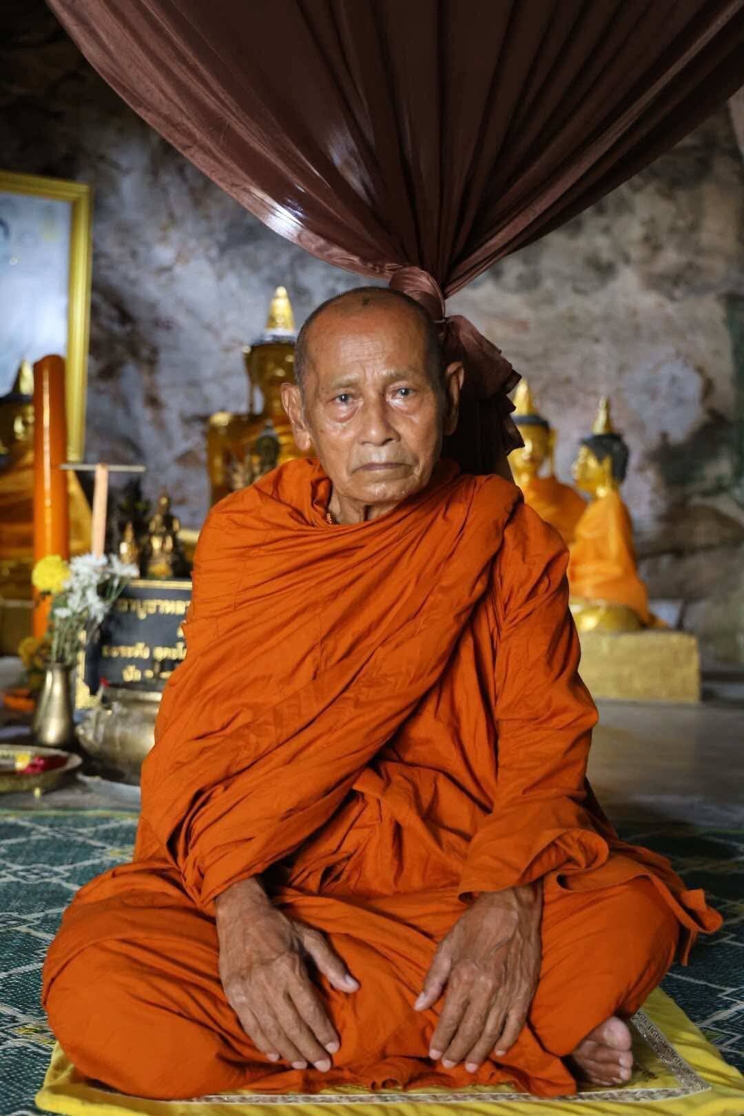Elder Thai man in orange robes seated in a temple interior holding blessed Thai Phan-Thai amulet for luck