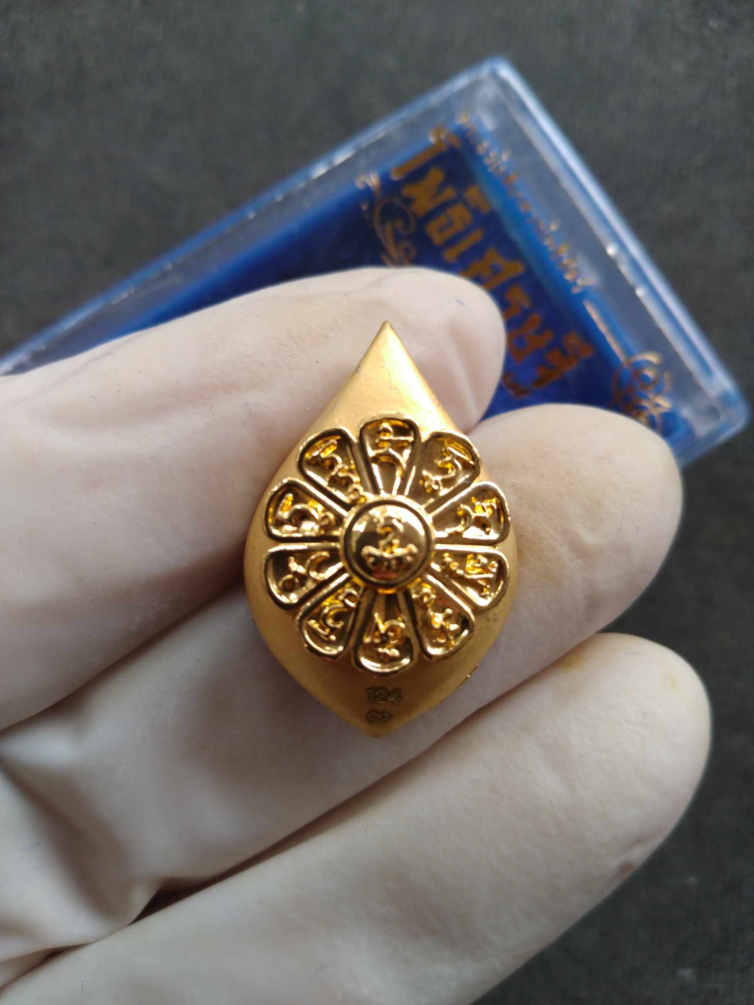 Gold teardrop amulet with floral wheel motif held by gloved hand Avalokiteshvara Bodhisattva Phothi Setthi Edition