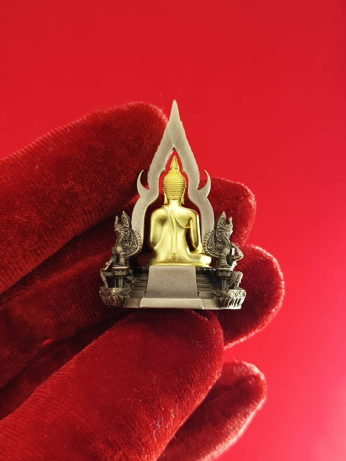 Phra Buddha Chinnarat amulet on a decorative base with intricate details held in a red glove