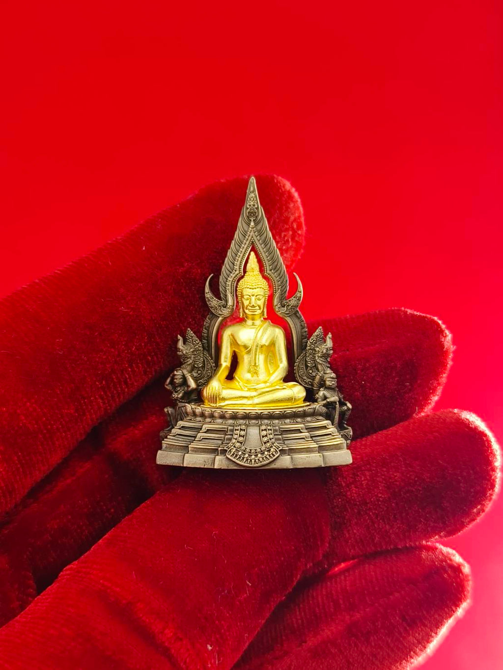 Phra Buddha Chinnarat amulet in a detailed design held in a gloved hand against a red background