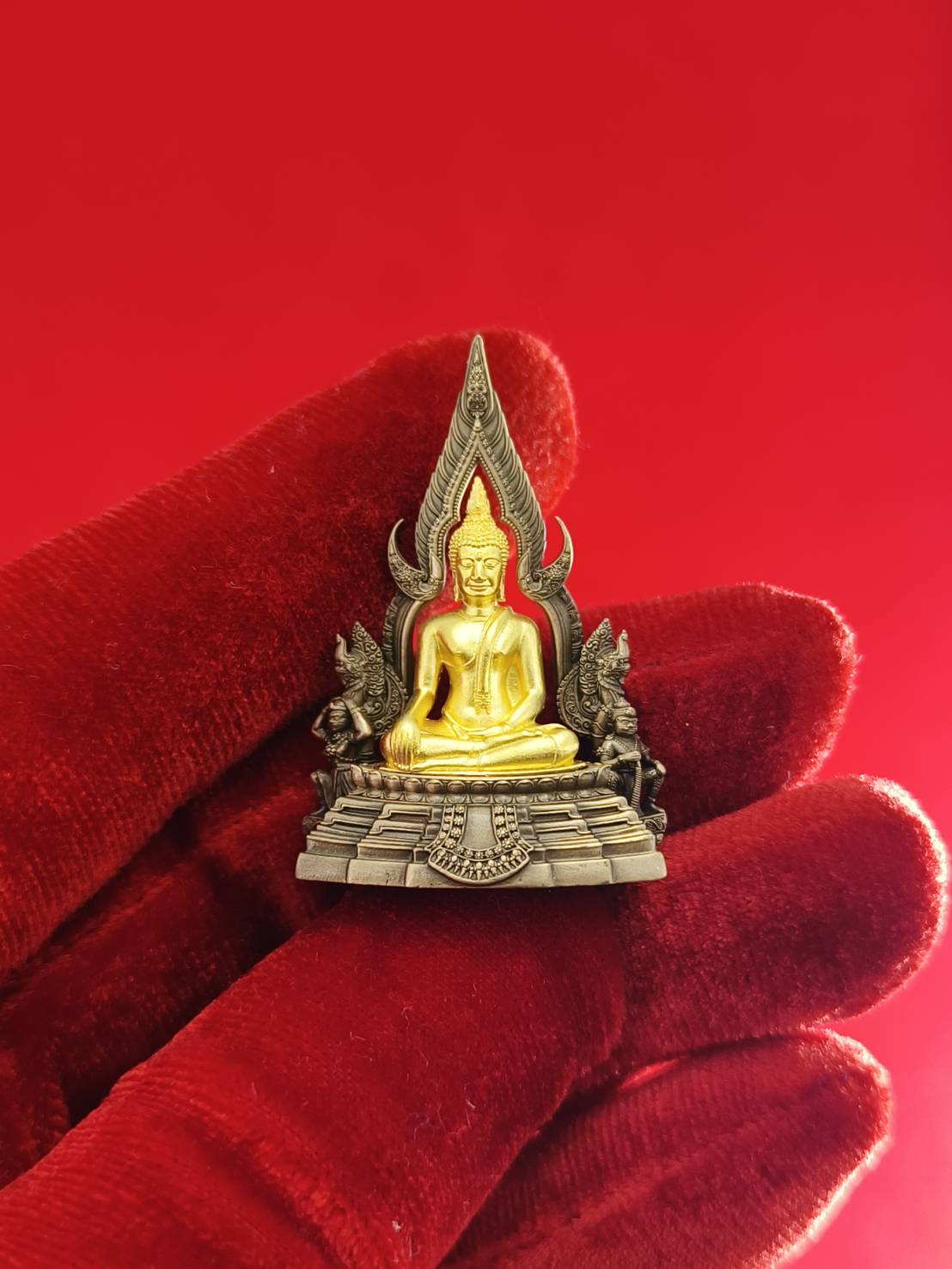 Phra Buddha Chinnarat amulet in gold and silver colors, held in a hand with a red background