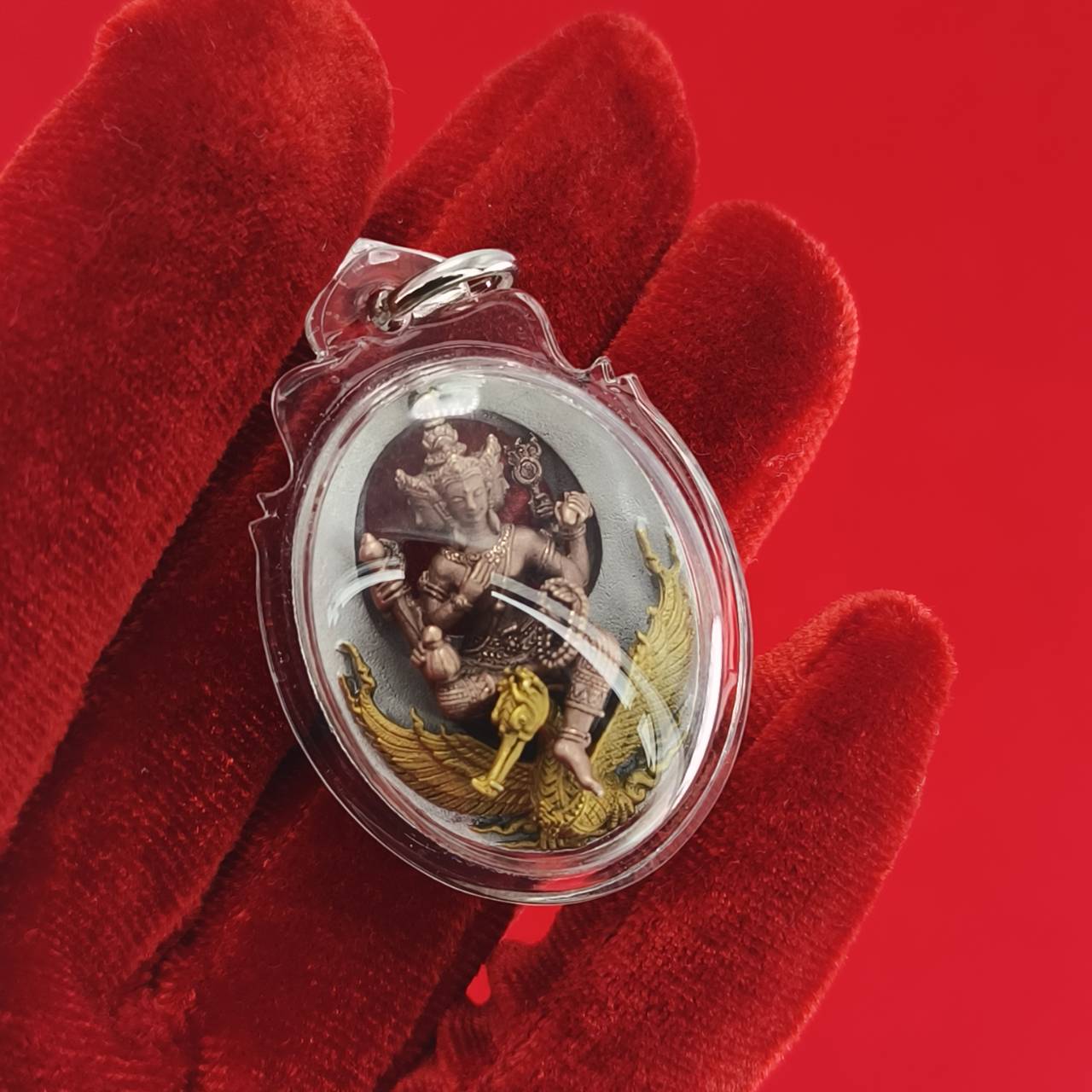 Phra Phrom Thai amulet in a clear pendant held by a red-gloved hand, symbolizing protection and blessings