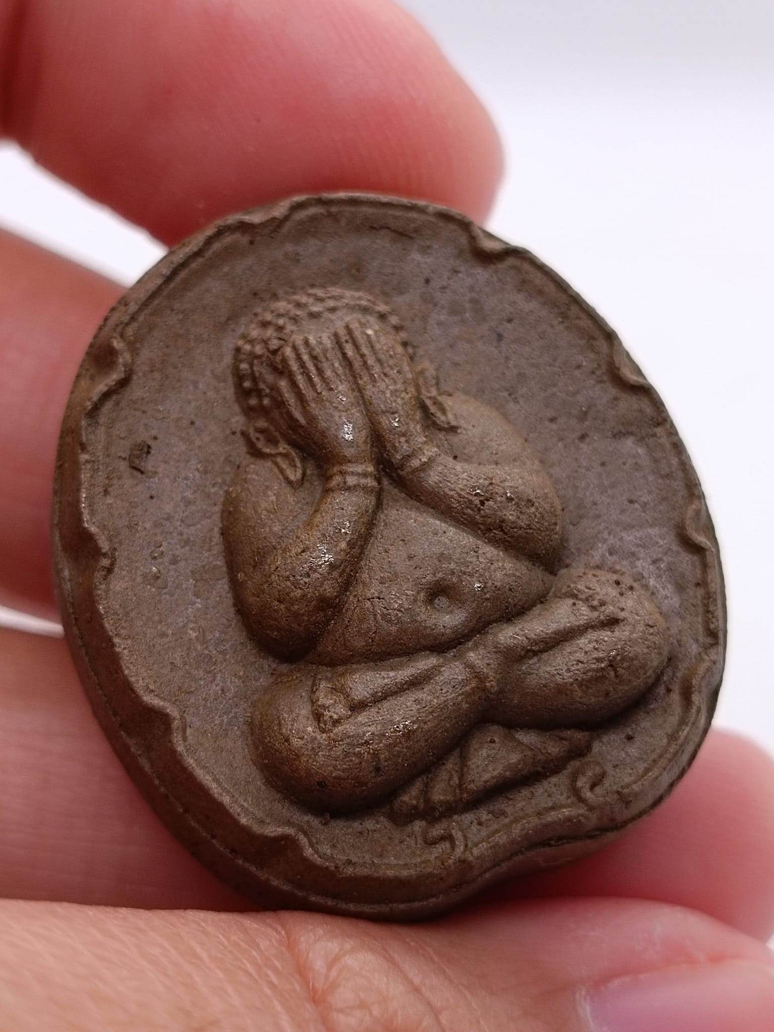 Phra Pidta wealth amulet depicted in textured clay with a seated figure covering its eyes