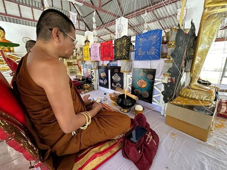 Monk blessing a Phra Rahu amulet for luck surrounded by decorative religious items