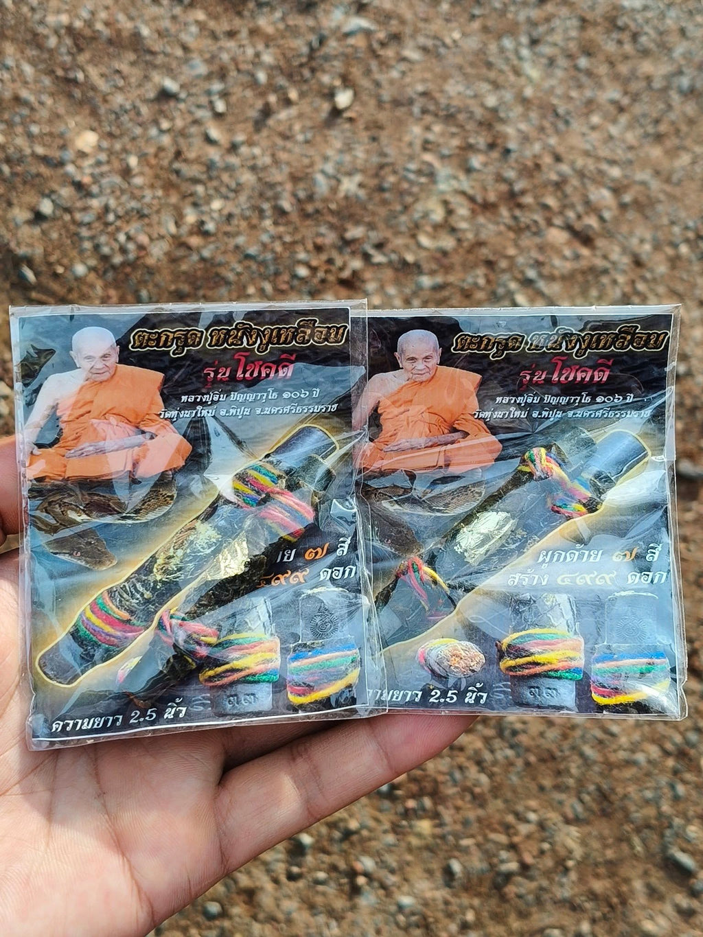 Hand holding packaged Buddhist amulet cards showing monk photo and authentic Thai python skin talisman wrapped in colorful cords