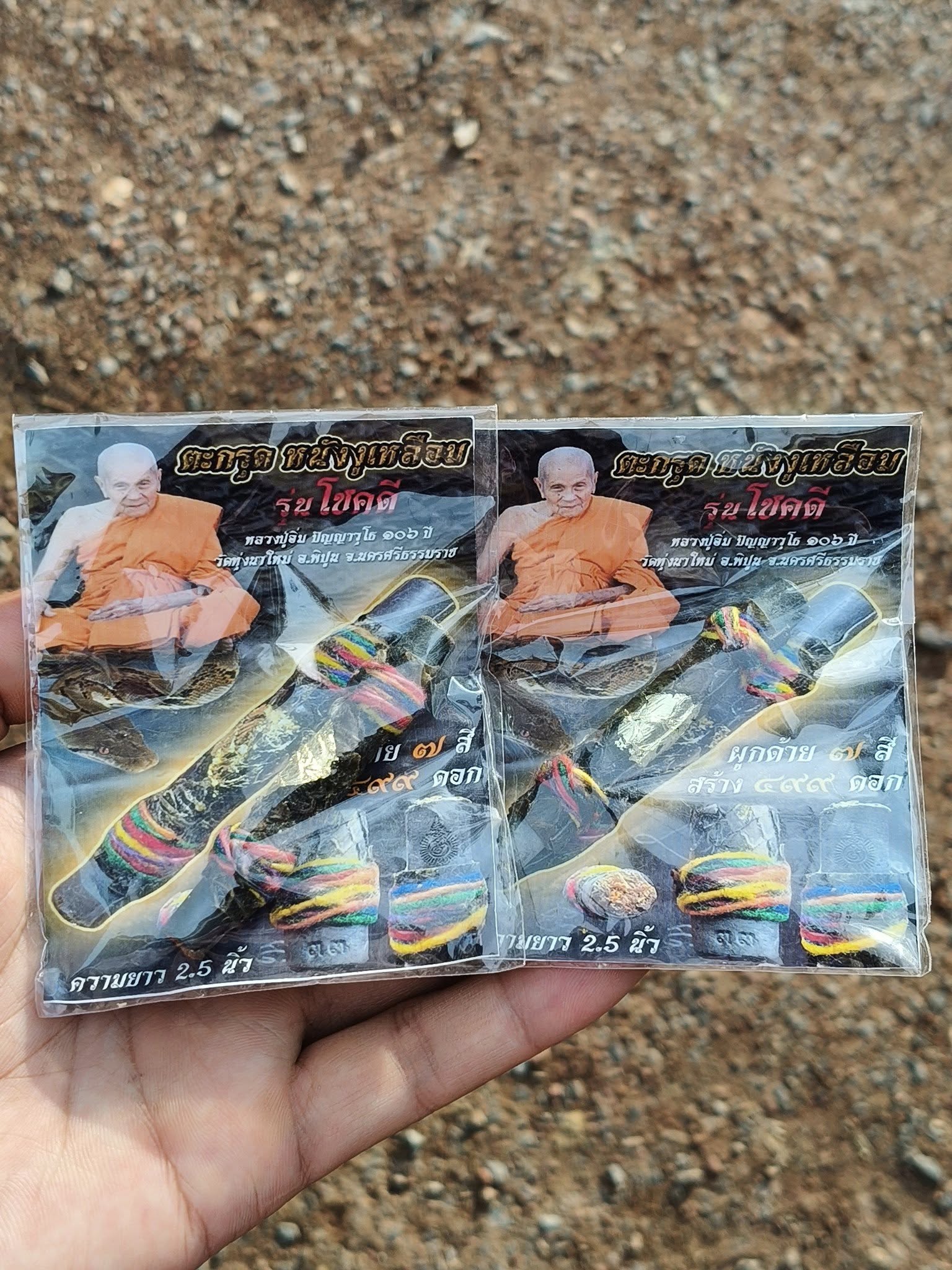 Hand holding packaged Buddhist amulet cards showing monk photo and authentic Thai python skin talisman wrapped in colorful cords