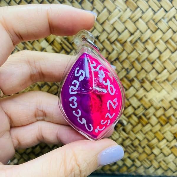 Hand holding Thai golden face charm pendant with red and purple colors and Thai script on woven background
