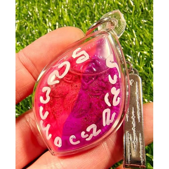 Hand holding Thai golden face charm pendant in pink and purple encased in clear plastic with white script