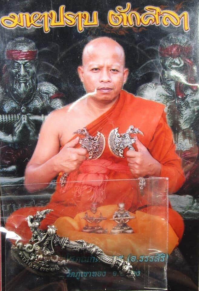 Thai monk holding two ornate axe amulets with small statues and display case Great Axe Amulet Maha Prap